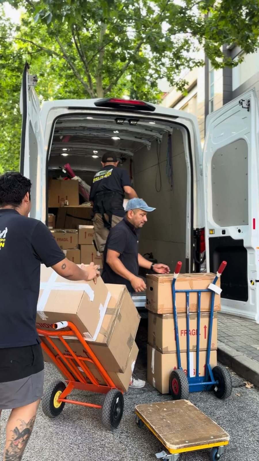 Tres operarios cargan cajas en la furgoneta con dos carretillas