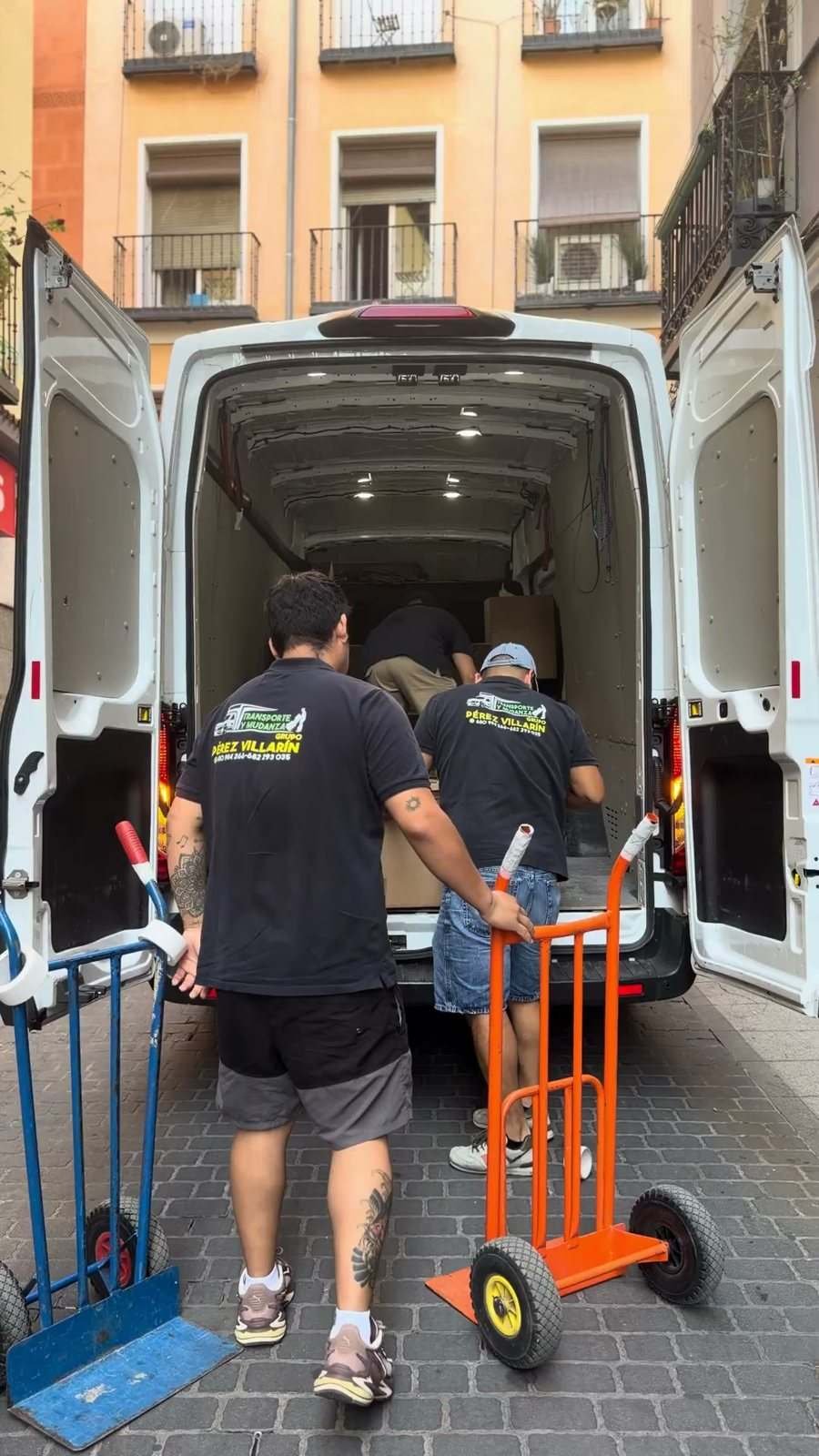 Two movers in Pérez Villarín T-shirts load a white van with hand trucks on a street of old façades
